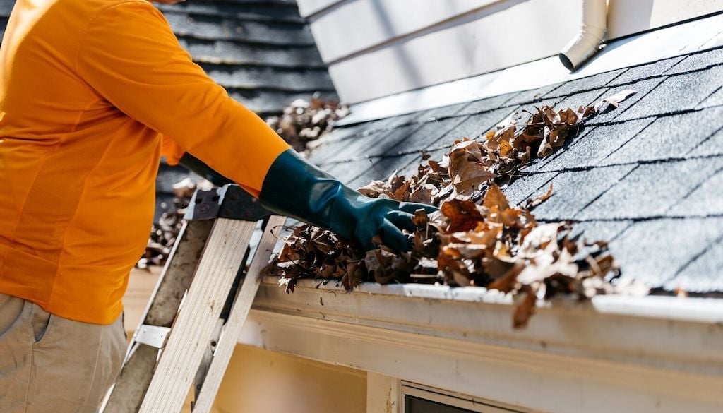 Worker in orange shirt and blue gloves cleaning debris from roof gutter during home maintenance