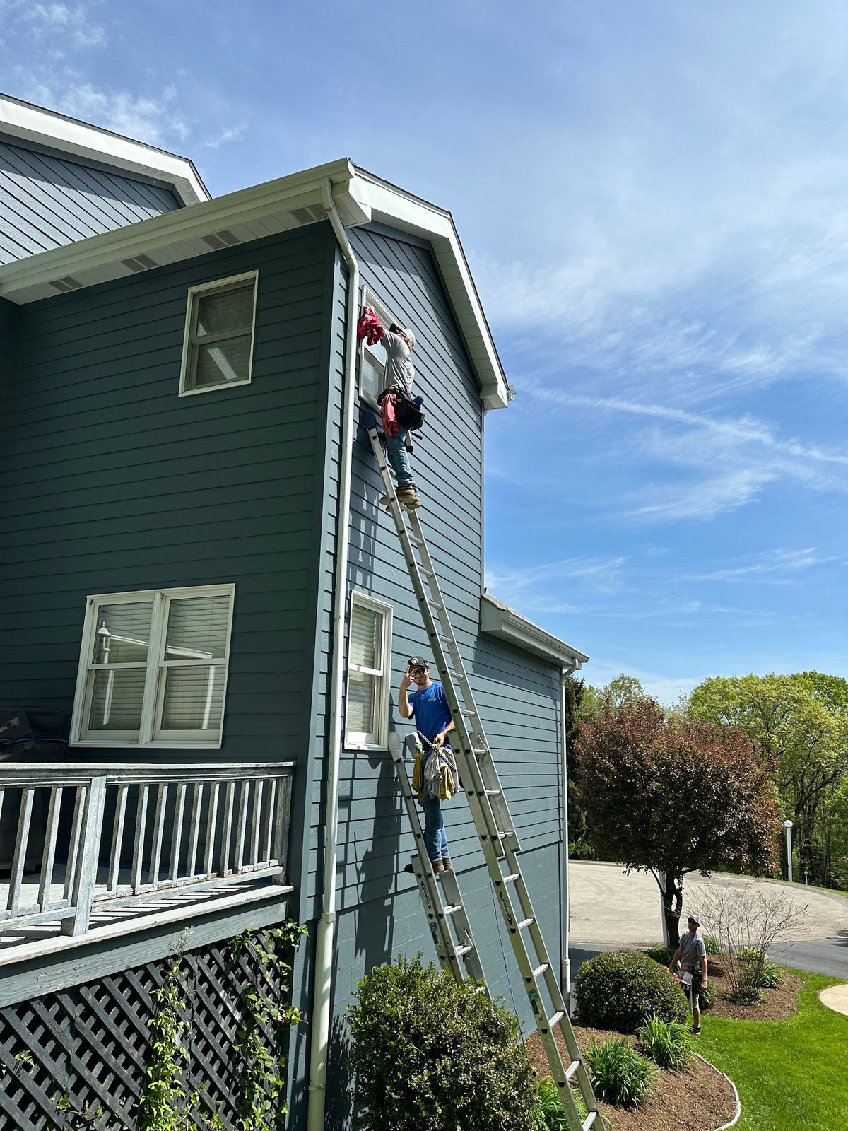 Two workers on extension ladders cleaning exterior siding and gutters of a two-story green and white house on a sunny day